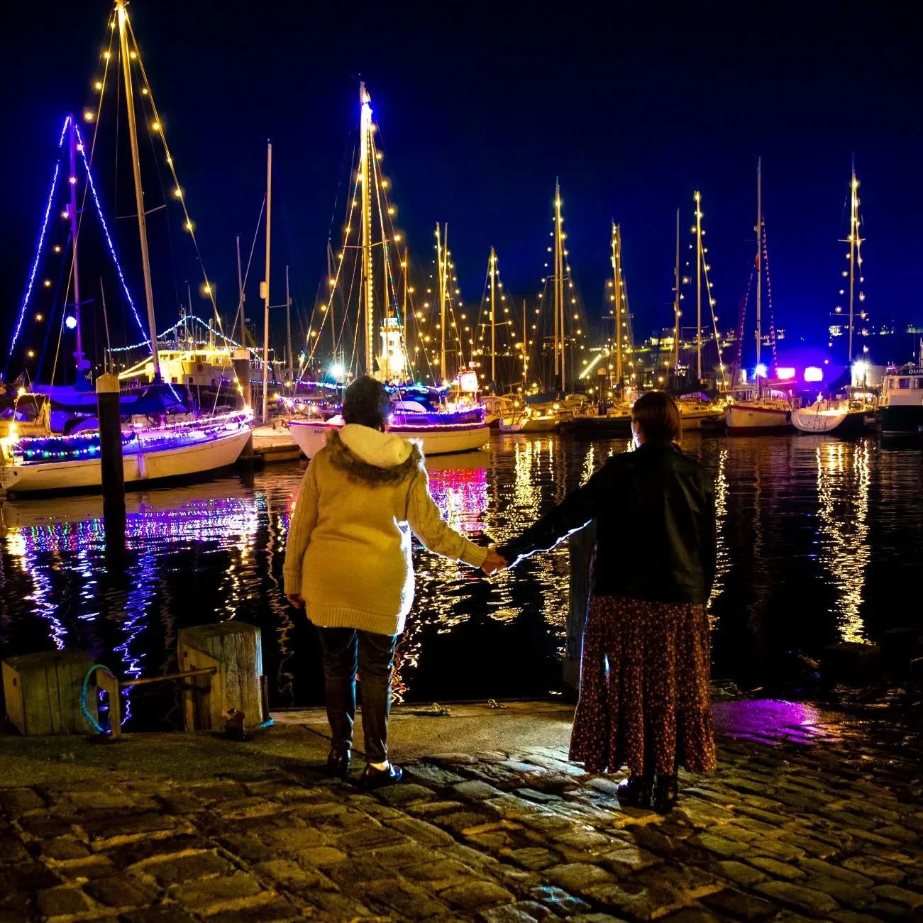 Two ladies hold hands in front of illuminated boats at Scarborough Harbour