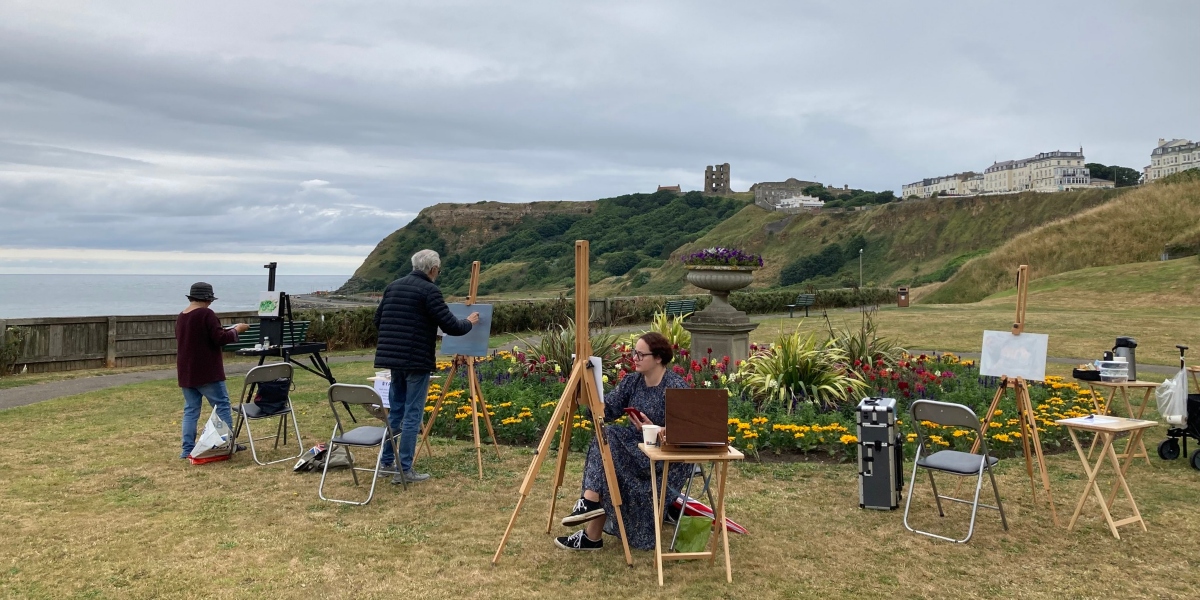 An image of artists sat on the cliff tops painting an image of Scarborough Castle.